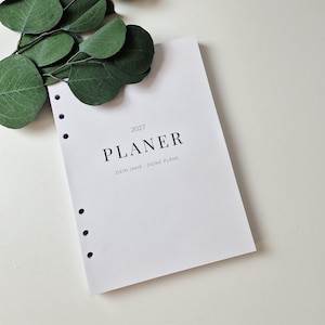 Op de afbeelding: Een wit papieren planner-inlegvel met zes ponsgaten langs de linkerrand. De omslag bevat de tekst 2027 PLANER en DEIN JAHR - DEINE PLÄNE in zwarte serif- en schreefloze lettertypes. Een takje met groene eucalyptusbladeren ligt linksboven.