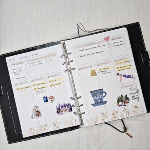 May include: Open black planner with a weekly layout. The pages are filled with handwritten notes, stickers, and decorative elements. The planner includes sections for tasks, trackers, and daily entries. The text "COLLECT moments not things" is visible.