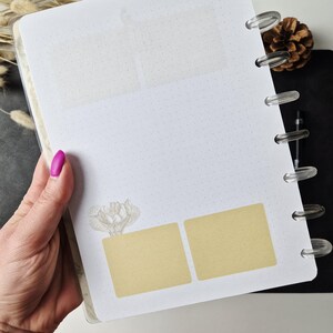 May include: A white disc-bound planner page featuring a light gray dot grid pattern, a translucent rectangular header section, and two solid light tan rectangular adhesive notes positioned above a small floral illustration.