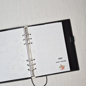 May include: Open black leather binder with white pages. The left page has a floral design, while the right page displays a 2027 calendar. The binder is open, revealing the metal ring mechanism and a black strap.
