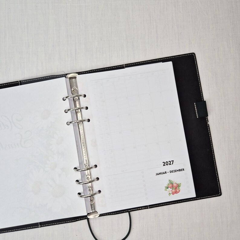 May include: Open black leather binder with white pages. The left page has a floral design, while the right page displays a 2027 calendar. The binder is open, revealing the metal ring mechanism and a black strap.