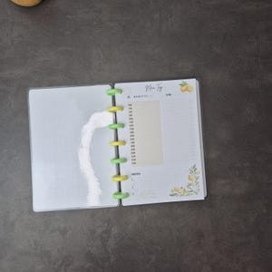 May include: Open planner with a clear plastic cover, featuring a white interior with a grid pattern on the left page. The right page has a time schedule and the text "Men Tyg" with lemon illustrations. The planner is held together by green and yellow disc binding.