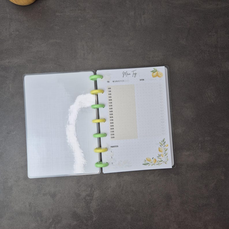 May include: Open planner with a clear plastic cover, featuring a white interior with a grid pattern on the left page. The right page has a time schedule and the text "Men Tyg" with lemon illustrations. The planner is held together by green and yellow disc binding.