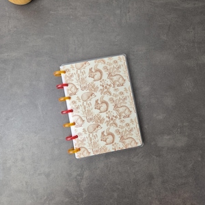 May include: A small notebook with a clear plastic cover and colorful binding rings. The cover features a repeating pattern of squirrels, rabbits, butterflies, and plants in shades of brown and cream. The binding rings alternate between red and yellow.