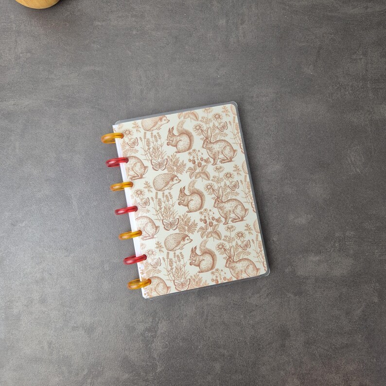 May include: A small notebook with a clear plastic cover and colorful binding rings. The cover features a repeating pattern of squirrels, rabbits, butterflies, and plants in shades of brown and cream. The binding rings alternate between red and yellow.