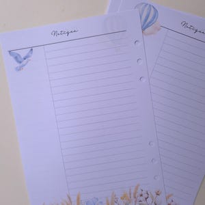 May include: Two lined paper sheets with watercolor illustrations. One sheet features a blue bird, the other a hot air balloon. Both have the word "Notizen" in script. The bottom of the sheets have floral and wheat illustrations.
