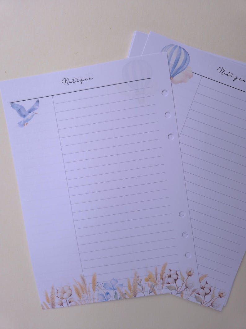May include: Two lined paper sheets with watercolor illustrations. One sheet features a blue bird, the other a hot air balloon. Both have the word "Notizen" in script. The bottom of the sheets have floral and wheat illustrations.