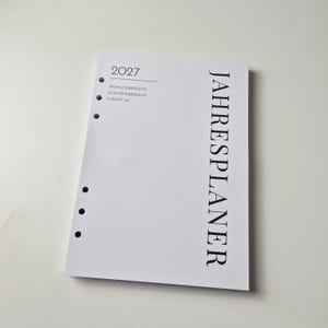 K&ouml;nnte beinhalten: Eine wei&szlig;e A5-Planer-Einlage f&uuml;r das Jahr 2027 mit sechs gelochten L&ouml;chern an der linken Seite. Der Text lautet 2027, MONATS&Uuml;BERSICHT, WOCHEN&Uuml;BERSICHT, FORMAT A5 und JAHRESPLANER in schwarzer Schrift. Das Design ist schlicht mit schwarzem Text.