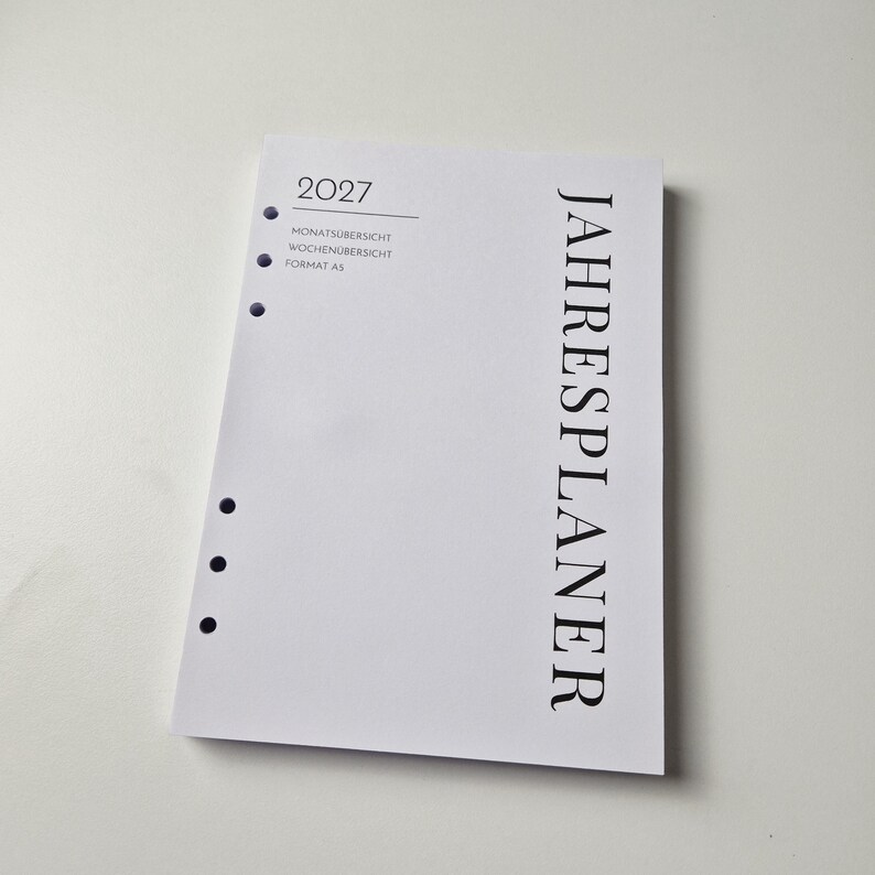 K&ouml;nnte beinhalten: Eine wei&szlig;e A5-Planer-Einlage f&uuml;r das Jahr 2027 mit sechs gelochten L&ouml;chern an der linken Seite. Der Text lautet 2027, MONATS&Uuml;BERSICHT, WOCHEN&Uuml;BERSICHT, FORMAT A5 und JAHRESPLANER in schwarzer Schrift. Das Design ist schlicht mit schwarzem Text.