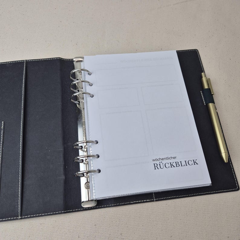 May include: A black leather-like binder with a six-ring metal mechanism holding white paper inserts. The paper features a grid layout and the German text w&ouml;chentlicher R&Uuml;CKBLICK. A gold-toned metal pen is secured in a loop on the right side.