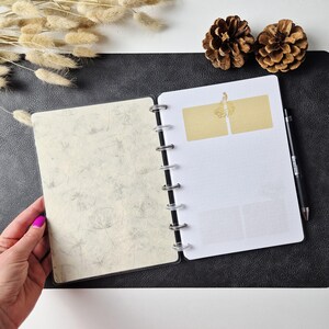 May include: A disc-bound planner with a cream-colored cover featuring a gray dandelion pattern. The interior white pages include two gold rectangular labels at the top and a black pen resting along the right side of the binding rings.