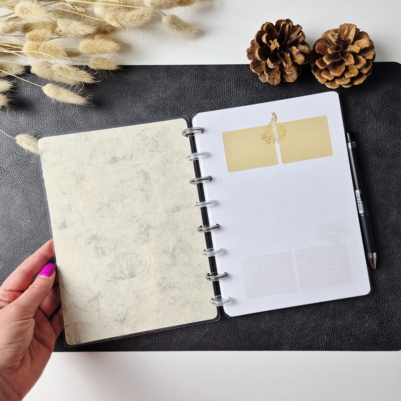 May include: A disc-bound planner with a cream-colored cover featuring a gray dandelion pattern. The interior white pages include two gold rectangular labels at the top and a black pen resting along the right side of the binding rings.