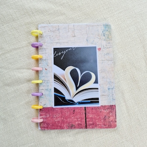Peut inclure: Un journal fait à la main avec une couverture décorative présentant une photographie d'un livre avec des pages pliées en forme de cœur. Le journal a un fond à motifs rouges et blancs et est relié avec des anneaux en plastique colorés. Le mot "lesejour" est écrit sur la photographie.
