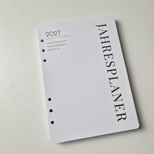 K&ouml;nnte beinhalten: Ein wei&szlig;er A5-Planer-Einleger f&uuml;r das Jahr 2027 mit schwarzem Text. Das Deckblatt zeigt vertikal das Wort JAHRESPLANER, sowie 2027, MONATS&Uuml;BERSICHT, WOCHEN&Uuml;BERSICHT und FORMAT A5 auf der linken Seite. Es verf&uuml;gt &uuml;ber sechs runde Ringbuchlocher.