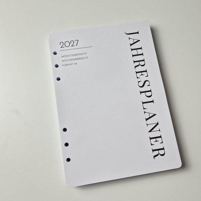 K&ouml;nnte beinhalten: Ein wei&szlig;er A5-Planer-Einleger f&uuml;r das Jahr 2027 mit schwarzem Text. Das Deckblatt zeigt vertikal das Wort JAHRESPLANER, sowie 2027, MONATS&Uuml;BERSICHT, WOCHEN&Uuml;BERSICHT und FORMAT A5 auf der linken Seite. Es verf&uuml;gt &uuml;ber sechs runde Ringbuchlocher.
