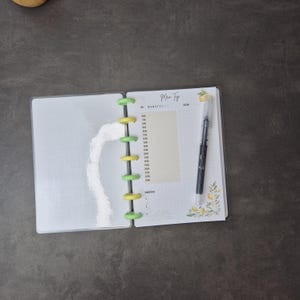 May include: Open planner with a clear plastic cover, featuring a daily schedule layout. The planner is held together with green and yellow disc binding. A black pen rests on the right page, next to a lemon and leaf design.