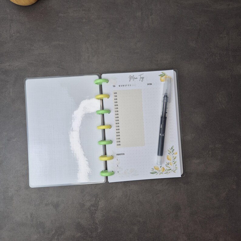 May include: Open planner with a clear plastic cover, featuring a daily schedule layout. The planner is held together with green and yellow disc binding. A black pen rests on the right page, next to a lemon and leaf design.