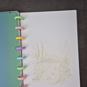 May include: Open planner with a gradient blue-green cover and dotted pages. The binding features colorful plastic discs. The right page has a soft illustration of a path with paw prints. A perfect tool for organizing notes and schedules.
