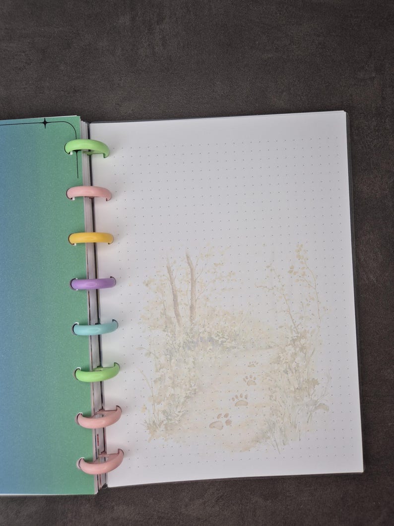 May include: Open planner with a gradient blue-green cover and dotted pages. The binding features colorful plastic discs. The right page has a soft illustration of a path with paw prints. A perfect tool for organizing notes and schedules.