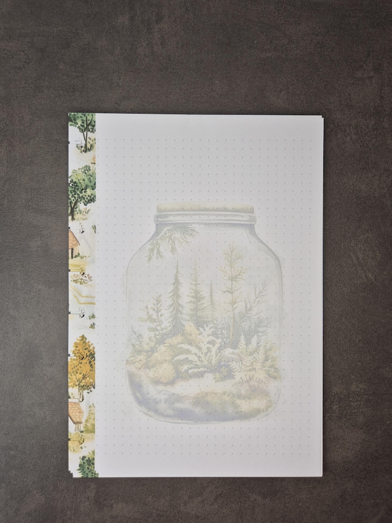 May include: A white notepad with a watercolor illustration of a terrarium inside a jar. The notepad has a dotted grid pattern and a decorative border with trees and houses. The jar contains trees and ferns.