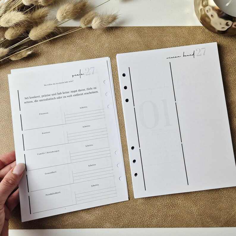 May include: Two white planner pages with black text and lines. The left page features sections for goals like Finanzen, Karriere, Familie, and Gesundheit with the text 'Sei konkret, pr&auml;zise und hab keine Angst davor'. The right page is titled 'vision board 27'.