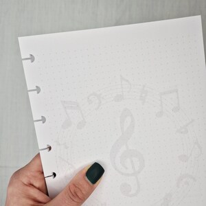 May include: White paper with a dotted grid and a faint musical note design. The paper has a series of holes on the left side. A hand with dark green nail polish holds the paper.