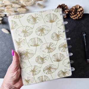 May include: A disc-bound notebook featuring a cream-colored cover with a repeating pattern of gold-toned, fan-shaped botanical illustrations. The notebook has clear plastic discs along the side and a transparent protective cover.