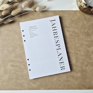 May include: A white planner insert page with six punched holes along the left side. The text JAHRESPLANER is printed vertically in large black letters. The page includes the year 2027 and a list of sections including ZIELE, KONTAKTE, and NOTIZEN.