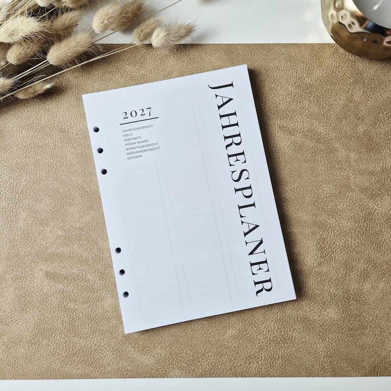 May include: A white planner insert page with six punched holes along the left side. The text JAHRESPLANER is printed vertically in large black letters. The page includes the year 2027 and a list of sections including ZIELE, KONTAKTE, and NOTIZEN.