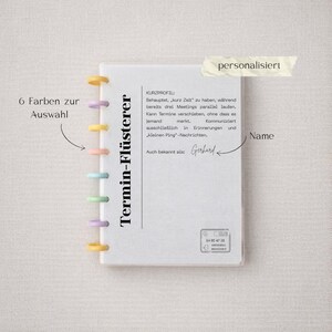 Op de afbeelding: Een wit notitieboek met de tekst "Termin-Flüsterer" en de naam "Gerhard". Het notitieboek heeft kleurrijke ringen en de tekst "6 Farben zur Auswahl". Een sticker leest "gepersonaliseerd".