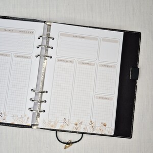 May include: Open planner with a black cover and silver rings. The pages are white with a weekly layout, including sections for tasks and a tracker. Floral design at the bottom of the pages. The planner is open to a weekly spread.
