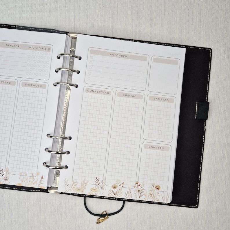 May include: Open planner with a black cover and silver rings. The pages are white with a weekly layout, including sections for tasks and a tracker. Floral design at the bottom of the pages. The planner is open to a weekly spread.