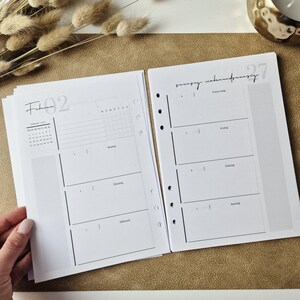 May include: A white paper planner insert with black ink, featuring a monthly calendar for February, a weekly schedule layout with days of the week in German, and punched holes along the side for binding. The pages include grid sections.