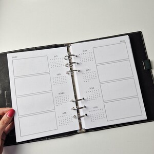 K&ouml;nnte beinhalten: Ein schwarzer Lederordner mit einer Sechs-Ring-Mechanik aus Metall und wei&szlig;en Einlagen. Die Seiten zeigen einen Kalender f&uuml;r 2027 mit Monats&uuml;bersichten von Januar bis August sowie leeren rechteckigen Feldern f&uuml;r Notizen.