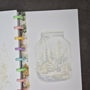 May include: Open notebook with a forest-in-a-jar illustration on a dotted page. The notebook has colorful plastic rings and a decorative strip with a nature-themed pattern. The illustration features trees and ferns inside a glass jar.
