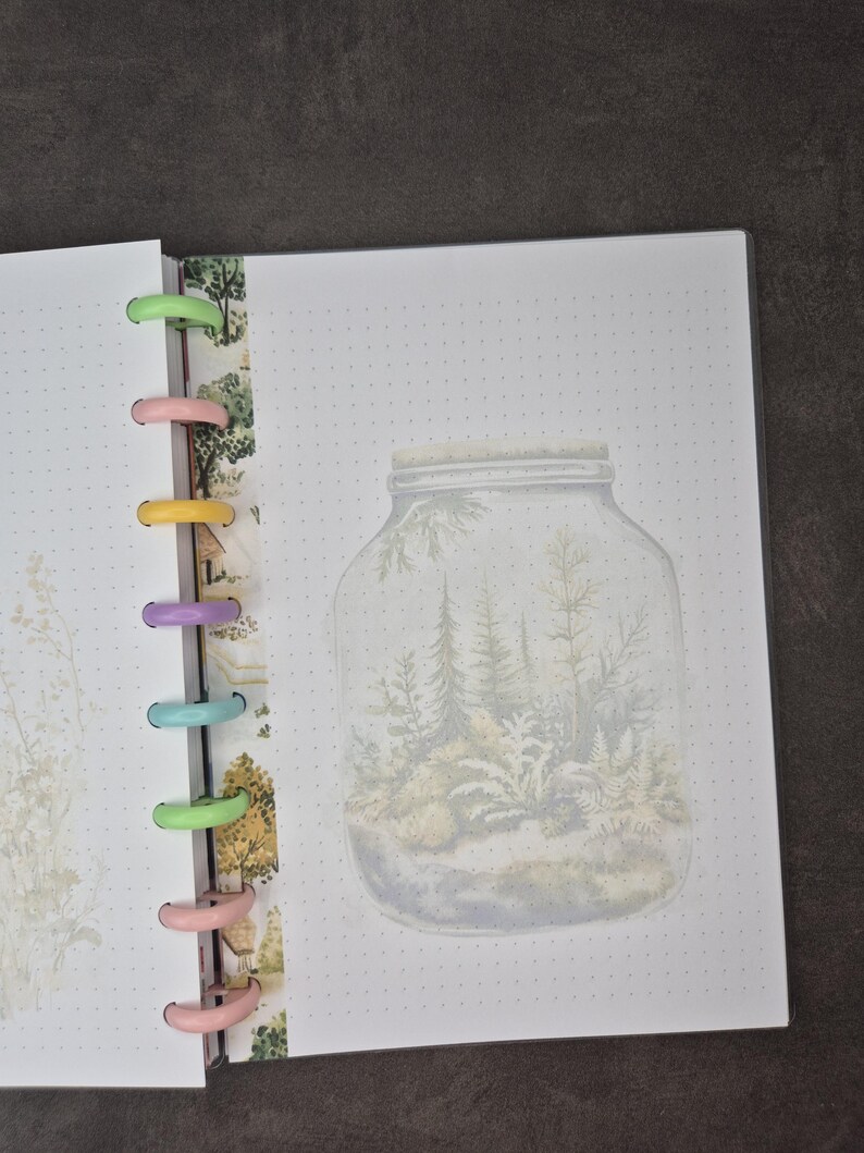 May include: Open notebook with a forest-in-a-jar illustration on a dotted page. The notebook has colorful plastic rings and a decorative strip with a nature-themed pattern. The illustration features trees and ferns inside a glass jar.