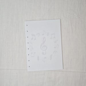 May include: A white sheet of paper with a musical theme. The paper has a faint pattern of musical notes and a treble clef. The sheet has several holes along the left edge, suggesting it is designed for a binder or ring binder.