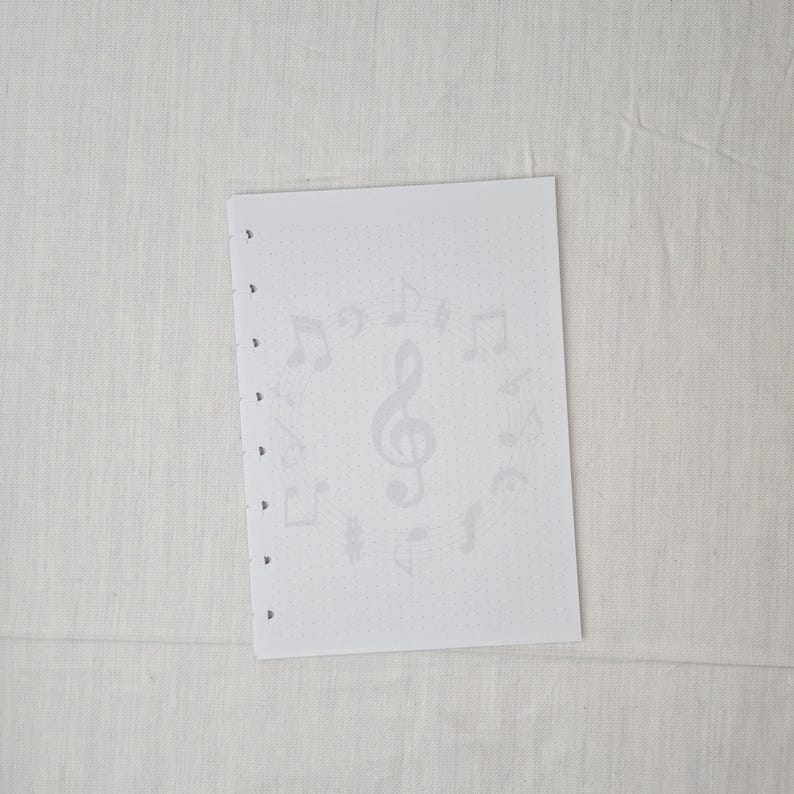 May include: A white sheet of paper with a musical theme. The paper has a faint pattern of musical notes and a treble clef. The sheet has several holes along the left edge, suggesting it is designed for a binder or ring binder.