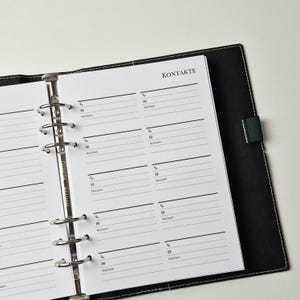K&ouml;nnte beinhalten: Ein schwarzer Ringbuchplaner mit wei&szlig;en Seiten und einem Kontaktlisten-Layout. Die Seiten&uuml;berschrift lautet KONTAKTE, mit Abschnitten f&uuml;r Telefonnummern, E-Mail-Adressen und Notizen. Der Metall-Ringbuchmechanismus verf&uuml;gt &uuml;ber sechs silberne Ringe.
