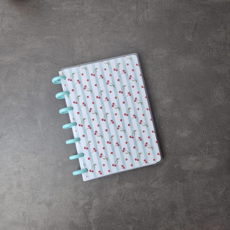 May include: A small notebook with a light blue cover and binding. The notebook's pages have a pattern of red cherries and green stems on a white background with light blue vertical stripes. The notebook is open.