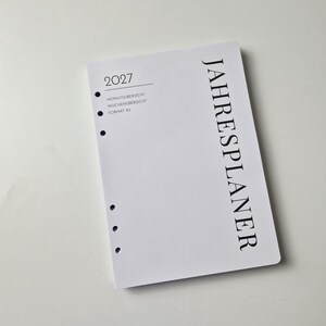 K&ouml;nnte beinhalten: Ein wei&szlig;es A5-Planerblatt f&uuml;r das Jahr 2027 mit schwarzem Text. Das Deckblatt zeigt das Wort JAHRESPLANER vertikal, sowie 2027, MONATS&Uuml;BERSICHT, WOCHEN&Uuml;BERSICHT und FORMAT A5 horizontal gedruckt. Es verf&uuml;gt &uuml;ber sechs kreisf&ouml;rmige L&ouml;cher am linken Rand.