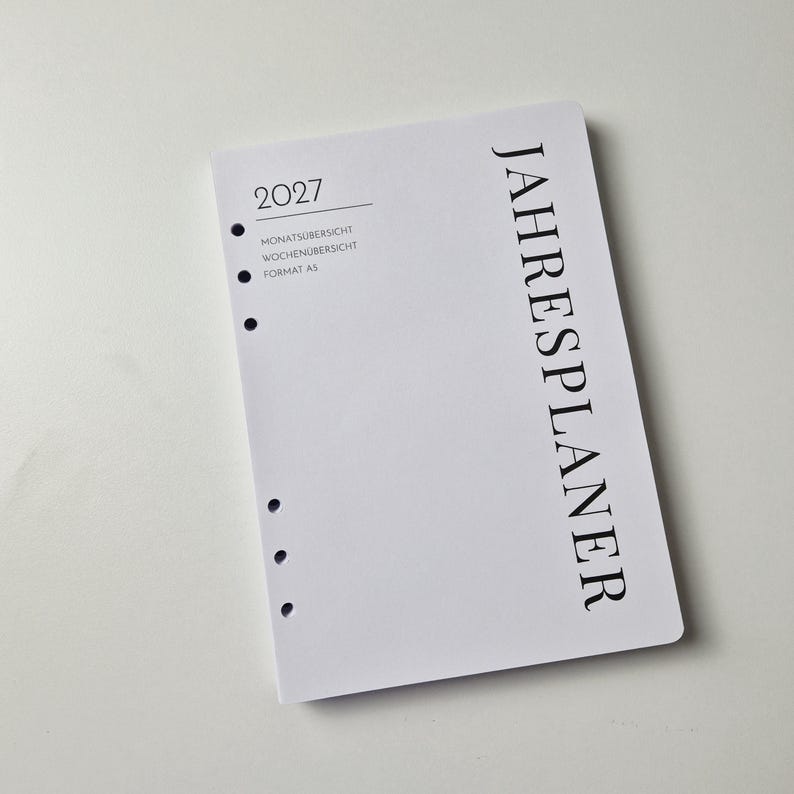 K&ouml;nnte beinhalten: Ein wei&szlig;es A5-Planerblatt f&uuml;r das Jahr 2027 mit schwarzem Text. Das Deckblatt zeigt das Wort JAHRESPLANER vertikal, sowie 2027, MONATS&Uuml;BERSICHT, WOCHEN&Uuml;BERSICHT und FORMAT A5 horizontal gedruckt. Es verf&uuml;gt &uuml;ber sechs kreisf&ouml;rmige L&ouml;cher am linken Rand.