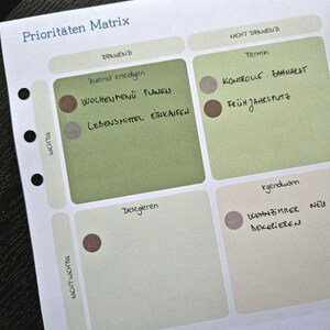 K&ouml;nnte beinhalten: Eine Planerseite mit dem Titel "Priorit&auml;ten Matrix" in Blau. Die Seite ist in Quadranten unterteilt und enth&auml;lt handschriftliche Notizen auf Deutsch, darunter "Zuerst erledigen" und "Wochenmen&uuml; planen".