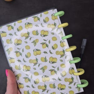 May include: A planner with a white cover featuring a pattern of yellow lemons and green leaves. The planner has a clear plastic cover and is held open with green and yellow rings. A pen is visible in the background.