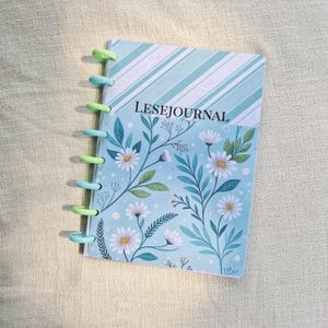 Peut inclure: Un petit journal à spirales avec une couverture bleu clair ornée d'un motif floral avec des marguerites blanches et des feuilles vertes. La partie supérieure présente des rayures diagonales. Le texte "LESEJOURNAL" est imprimé sur la couverture.