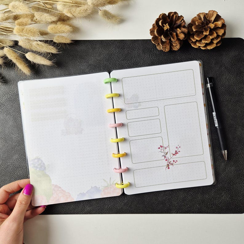May include: An open disc-bound planner with white pages featuring a dot grid pattern, rectangular text boxes, and floral illustrations. The planner has colourful plastic binding discs in yellow, green, and pink, and is accompanied by a black pen.