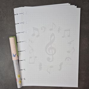 May include: A stack of white dot grid paper with a musical note design and a pen. The pen has a light pink cap with a butterfly and floral design. The paper has a faint musical note pattern.