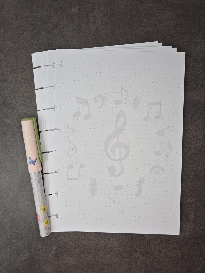 May include: A stack of white dot grid paper with a musical note design and a pen. The pen has a light pink cap with a butterfly and floral design. The paper has a faint musical note pattern.