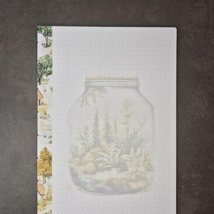 May include: A white notepad with a watercolor illustration of a terrarium inside a mason jar. The notepad has a dotted grid pattern and a decorative border with trees and houses. The background is a dark gray.