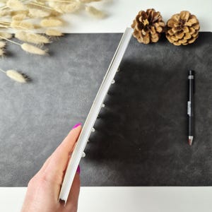 May include: A thin, white, rectangular notebook with a spiral binding, held vertically. The notebook features a clean, white spine and edges, showcasing its slim profile and simple construction for writing or note-taking purposes.